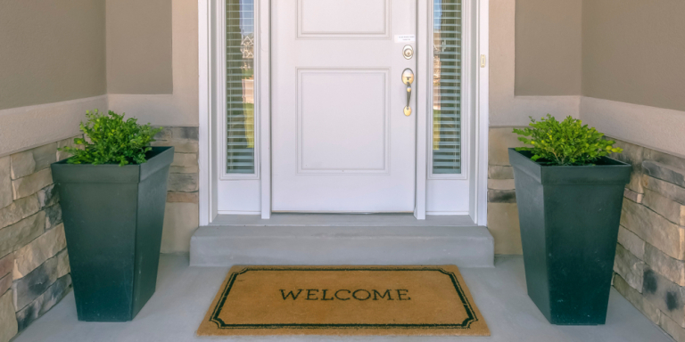Eine weiße Haustür, zwei Blumentöpfe und eine Fußmatte mit der Aufschrift "Welcome".